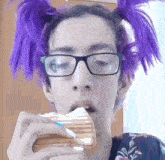 a girl with purple hair is eating a sandwich while wearing glasses .