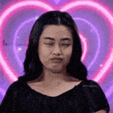 a woman is making a face with her eyes closed in front of a heart shaped neon sign .