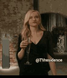 a woman in a black dress is holding a glass of wine in front of a brick wall ..