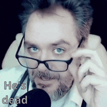 a man with glasses and a beard is talking into a microphone and the words he 's dead are above him