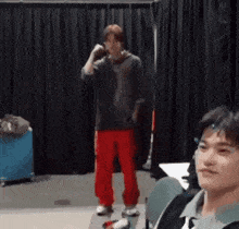a man in red pants is standing in front of a black curtain while another man looks on .