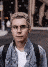 a young man wearing glasses and a denim jacket is standing in front of a building .