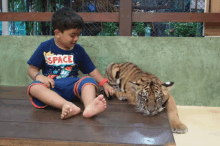 a young boy wearing a space shirt is sitting next to a tiger cub