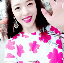 a woman in a pink and white floral shirt is smiling and waving her hand