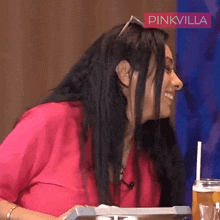a woman in a pink shirt is sitting at a table with a drink in front of her and a sign that says pinkvilla