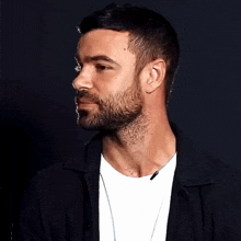 a man with a beard and a white t-shirt is wearing a black jacket and a necklace .