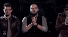 a man with a beard is praying with his hands folded in front of a group of people .