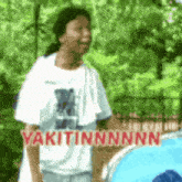 a man in a white shirt is standing in front of a fence with the words yakitinnnnn written on it .