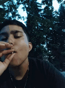 a man wearing a black shirt is smoking a cigarette with trees in the background