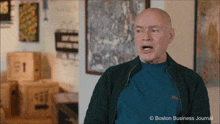 a bald man in a blue sweater stands in front of a boston business journal advertisement