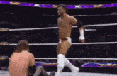 a wrestler is standing in the middle of a wrestling ring while another wrestler sits on the floor .