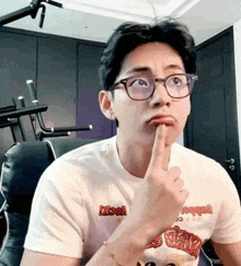 a man wearing glasses and a white t-shirt is making a funny face with his finger to his mouth .