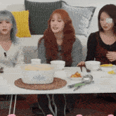 three women are sitting at a table with bowls and a casserole dish