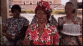 a woman wearing a red hat and a floral shirt is surrounded by other women holding plates of food .