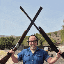 a man wearing headphones and glasses holds two guns crossed over his head