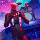 a man in a red suit is making a heart shape with his hands while standing in front of a drum set .