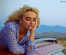 a woman with a cigarette in her mouth leans on a car