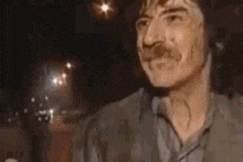 a man with a mustache is sitting at a table in a dark room and smiling .