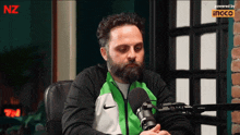 a man with a beard is sitting in front of a microphone with a nz logo in the background