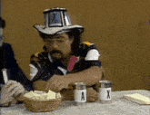 a man wearing a cowboy hat is sitting at a table with a basket of chips and two cans labeled a and x