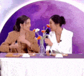 two women are sitting at a table with a vase of flowers in front of them