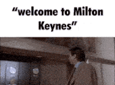 a man in a suit and tie is standing in front of an elevator with the words " welcome to milton keynes " above him
