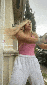 a woman in a pink tank top and white sweatpants is standing in front of a building holding her hair .
