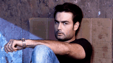 a man wearing a watch sits on a cardboard box against a wall