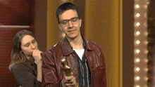 a man in a leather jacket is holding a trophy while a woman looks on behind him .