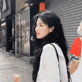a woman with long black hair is standing on a sidewalk next to a man wearing a face mask .