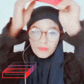a woman wearing a hijab and glasses holds a red object