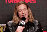 a man with long blonde hair is speaking into a microphone with the letters rt on it