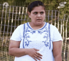 a woman in a white shirt with blue embroidery is standing in front of a fence and a sign that says wife swap