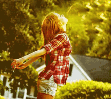 a woman in a plaid shirt and shorts stands in front of a house with her arms outstretched