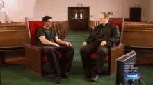 two men sit in red chairs in a church talking to each other with a travel channel monitor in front of them