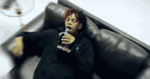 a man in a black hoodie is laying on a couch drinking from a bottle .