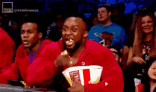 a man in a red jacket is eating popcorn while watching a wrestling match .