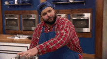 a man in a plaid shirt and blue apron is standing in a kitchen with a pot .