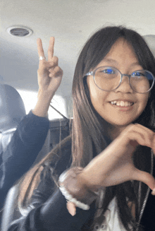 a girl wearing glasses is making a heart shape with her hands and giving a peace sign
