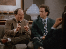 two men are sitting on a couch talking and one is pointing
