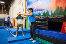 a woman in a blue shirt stands on a blue mat in front of a wall with a painting of mountains on it