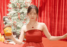 a woman in a red dress with a pearl necklace is holding a potted plant in front of a christmas tree