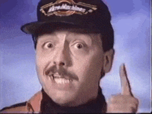 a man with a mustache is wearing a hat and pointing his finger .