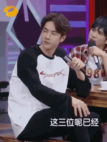 a man is sitting at a table holding a microphone while another man looks on .
