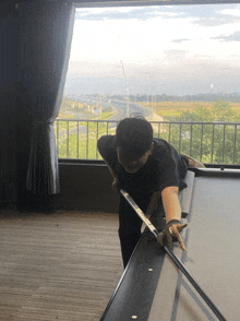 a man playing pool in front of a window with a highway in the background