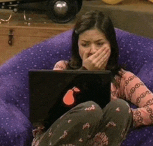 a woman sits in a purple chair covering her mouth while using an apple laptop