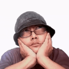 a man wearing a hat and glasses is sitting with his hands on his face .