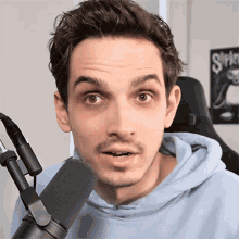 a man in a blue hoodie stands in front of a microphone with a slipknot poster in the background