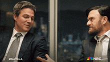 two men in suits and ties are having a conversation in front of a window with nbc written on the bottom
