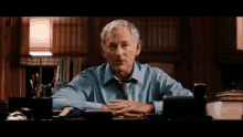 a man in a blue shirt and tie is sitting at a desk with his hands folded